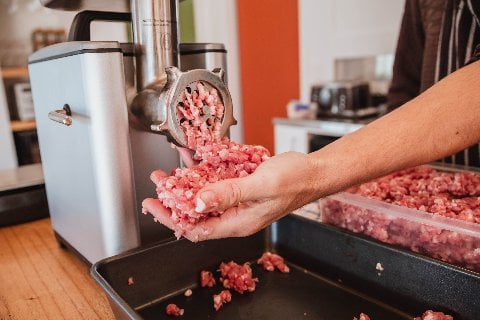 A person using a meat grinder to process raw meat, with freshly ground meat falling into their hand.