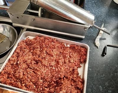 A tray filled with cuts of meat beside a sausage stuffing machine, suggesting someone is in the process of making homemade sausages.